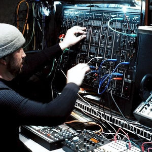 Producer in a beanie hat patching cables on a large modular synthesizer in a dimly lit studio