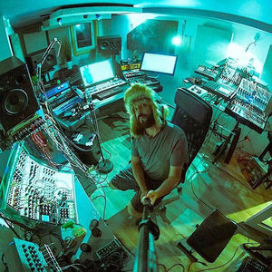 Producer smiling amid synthesizers and gear in the studio, the whole setup bathed in teal fisheye light
