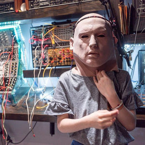 Person in a blank face mask standing before the Zombie Nation Eurorack modular, brightly lit with patch