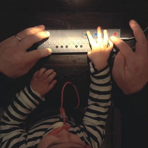 Adult and toddler hands playing a Teenage Engineering OP-Z together on a dark wooden kitchen table
