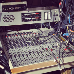 Vintage mixing console with multiple fader channels and outboard rack gear above, tape machine head stack