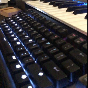 Blue-backlit mechanical keyboard on a studio desk with piano keys of a synthesizer visible behind