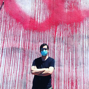 Person in a blue surgical mask and black top standing before a pink drip-paint graffiti mural outdoors