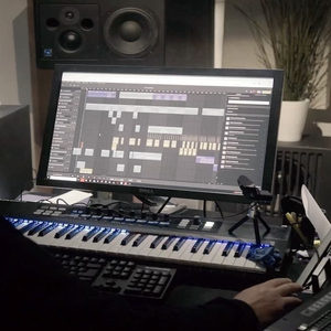 DAW arrangement open on a Dell monitor, blue-lit MIDI keyboard below and studio monitors above on a desk
