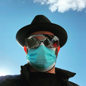 Florian Senfter outdoors in a black hat, mirrored visor sunglasses and teal surgical mask, blue sky