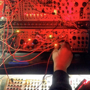 Hands patching a large modular synthesizer bathed in red light, with a step sequencer controller below