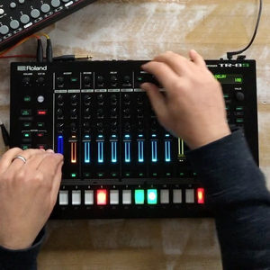 Overhead shot of two hands operating a Roland TR-8S drum machine, coloured step buttons and faders lit up