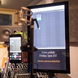 Studio monitor displaying a DAW and live collab event text, phone on a mic stand, mixing desk in front
