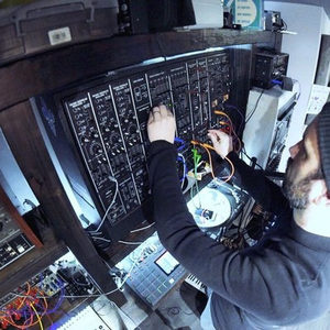 Overhead CCTV-style shot of two people at a large modular synthesizer, warm lighting and patch cables