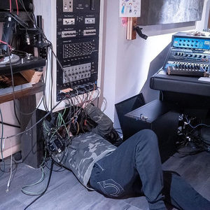 Person hunched in a corner of a studio surrounded by modular synthesizer racks and outboard gear