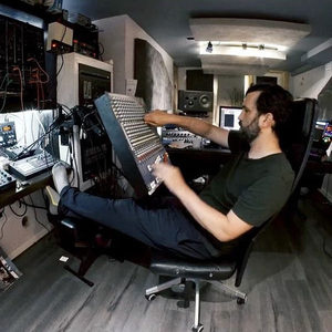 Producer reclined in a studio chair reaching across a large mixing console, modular racks on the left