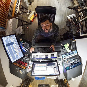 Producer at the studio desk seen from above, surrounded by synthesizers, a MIDI keyboard and dual screens