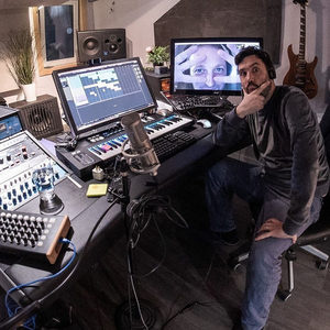 Producer at a studio desk with a condenser mic and DAW open, a video call face visible on a second monitor