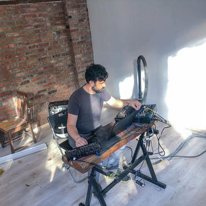 Producer at a trestle desk with a keyboard and Eurorack gear, bright ring light and brick wall behind