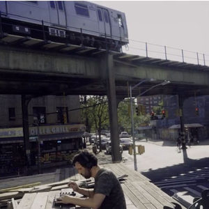 Producer at a keyboard and gear setup on a deck under a Bushwick elevated train, Deli and Grill sign behind