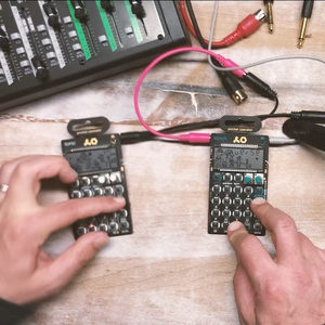 Two hands holding a pair of Teenage Engineering Pocket Operator devices side by side on a studio desk